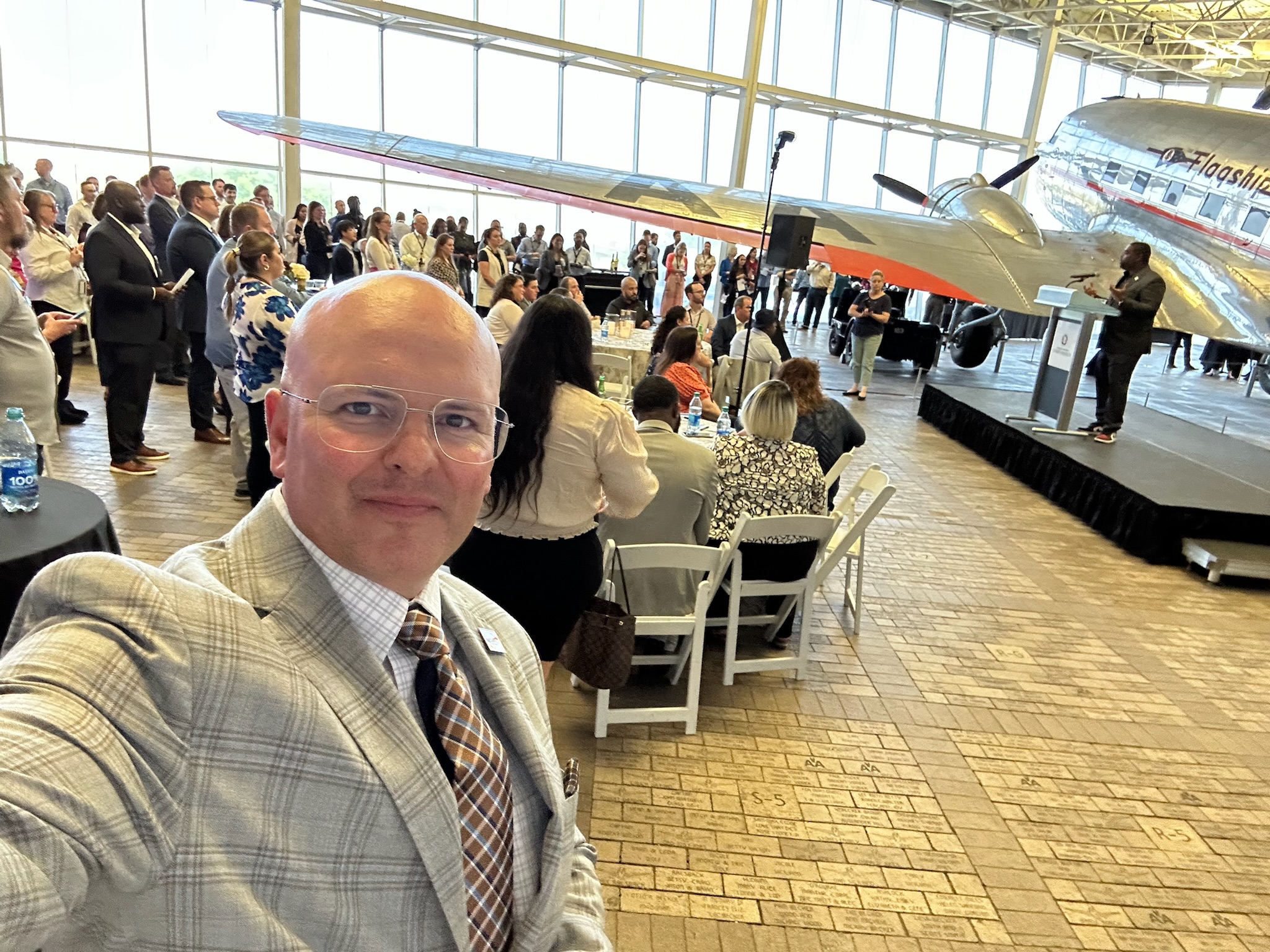 Robert Hausmann at a leadership gathering beside a historic aircraft, with attendees and a speaker on stage in the background.